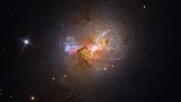 Dwarf starburst galaxy Henize 2-10 sparkles with young stars in this Hubble visible-light image. The bright region at the center, surrounded by pink clouds and dark dust lanes, indicates the location of the galaxy's massive black hole and active stellar nurseries.