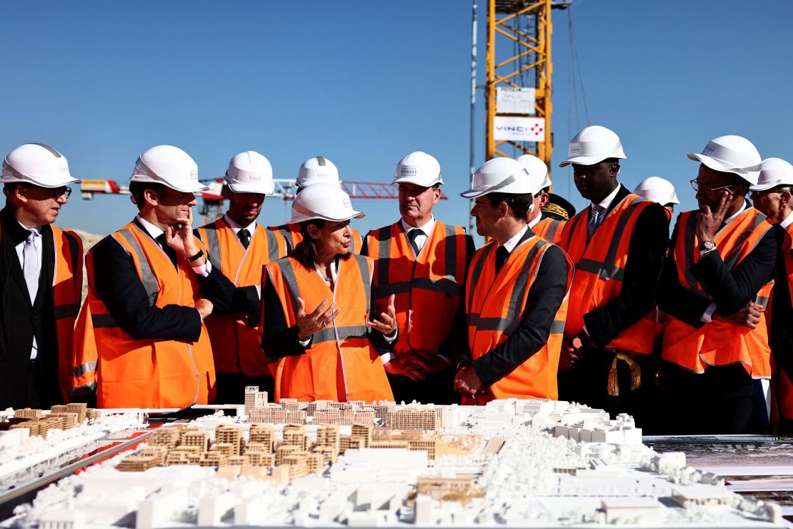 Paris Mayor Anne Hidalgo (C) speaks past French President Emmanuel Macron (second L) during a visit of the building site of the Olympic Village in Saint-Ouen on October 14, 2021.