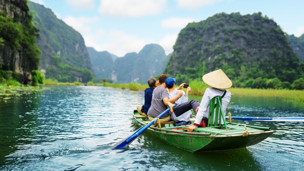 Tourists traveling in boat along the Ngo Dong River and taking picture of the Tam Coc, Ninh Binh, Vietnam. Rower using her feet to propel oars. Landscape formed by karst towers and rice fields.