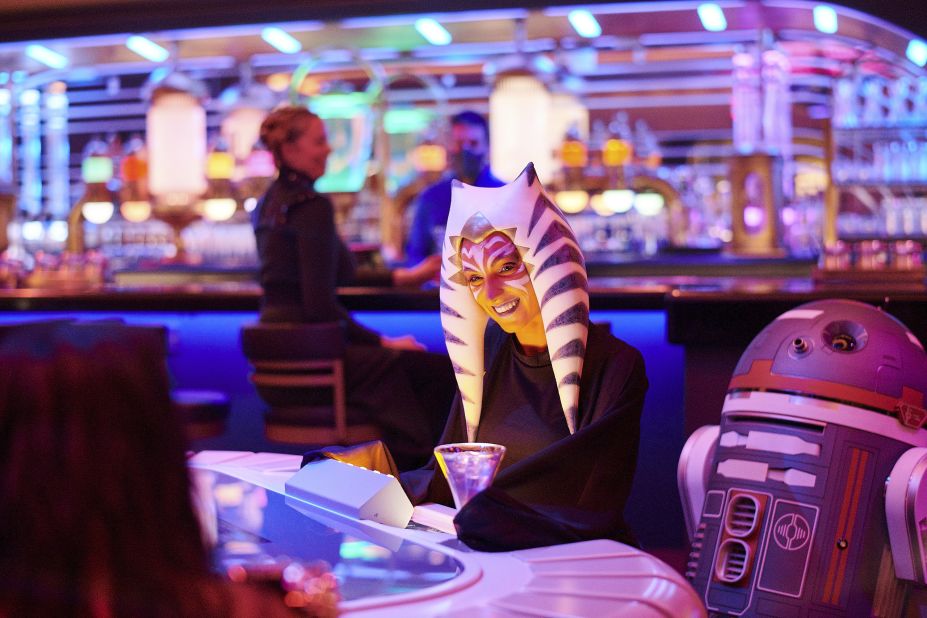 A passenger wearing a Togruta headpiece, similar to fan favorite character Ahsoka Tano, sits in the Sublight Lounge next to astromech droid SK-26O. Guests can purchase headpieces while onboard.