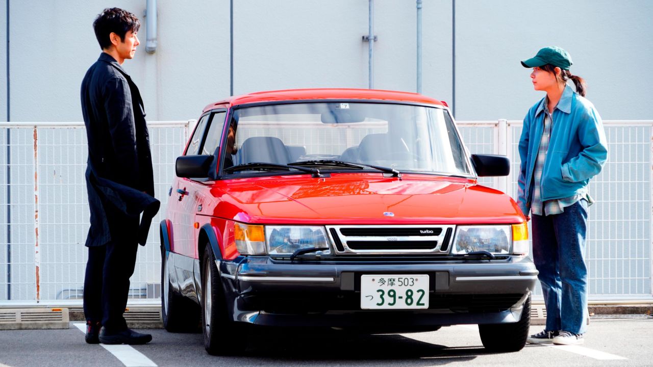 Hidetoshi Nishijima and Toko Miura as Yusuke Kafuku and Misaki Watari standing next to the red Saab 900 Turbo in Hamaguchi's "Drive My Car." 