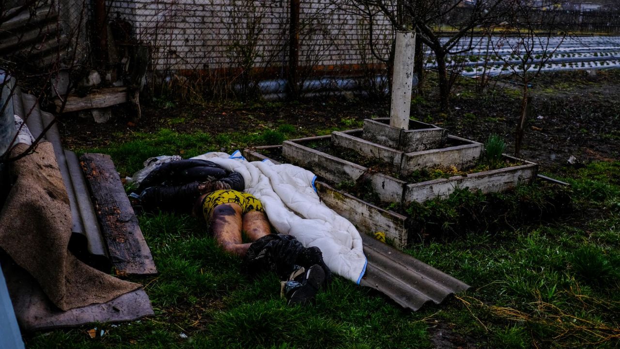 A man lays on the ground, hands tied behind his back, a gunshot wound on his head and a bullet casing next to his body.