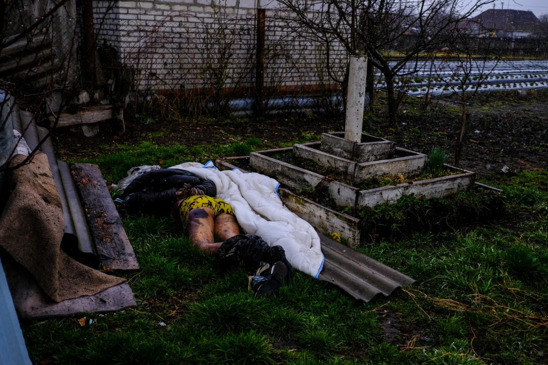 A man lays on the ground, hands tied behind his back, a gunshot wound on his head and a bullet casing next to his body.