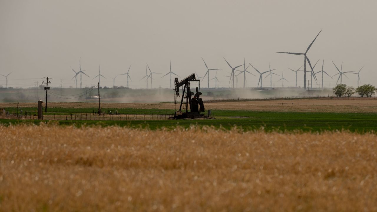 A single oil pump sits in a field in Weatherford, Oklahoma, as dozens of wind turbines spin in the background.