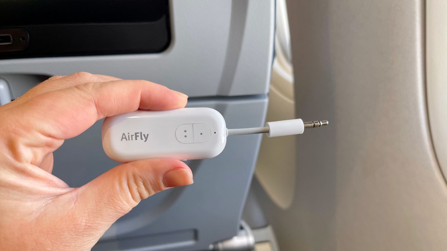 A photo of a hand holding a Twelve South AirFly Duo in front of an airplane seat