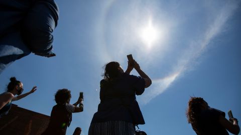 People look at a rainbow forming a circle around the sun as they mourn and pray on the street outside of Tops.