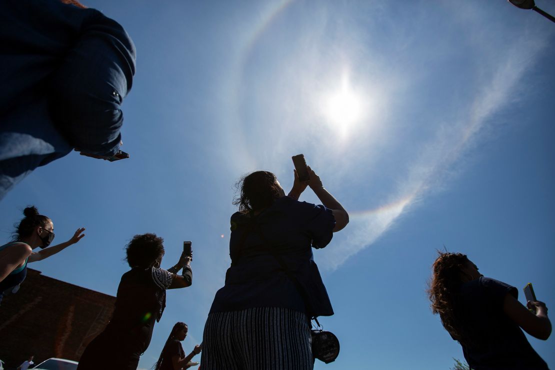 People look at a rainbow forming a circle around the sun as they mourn and pray on the street outside of Tops.