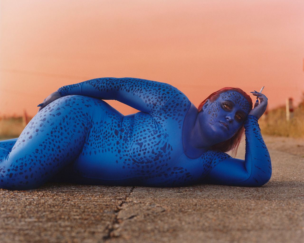Mystique, from "X-Men," is photographed lying on the ground, cigarette in hand. 