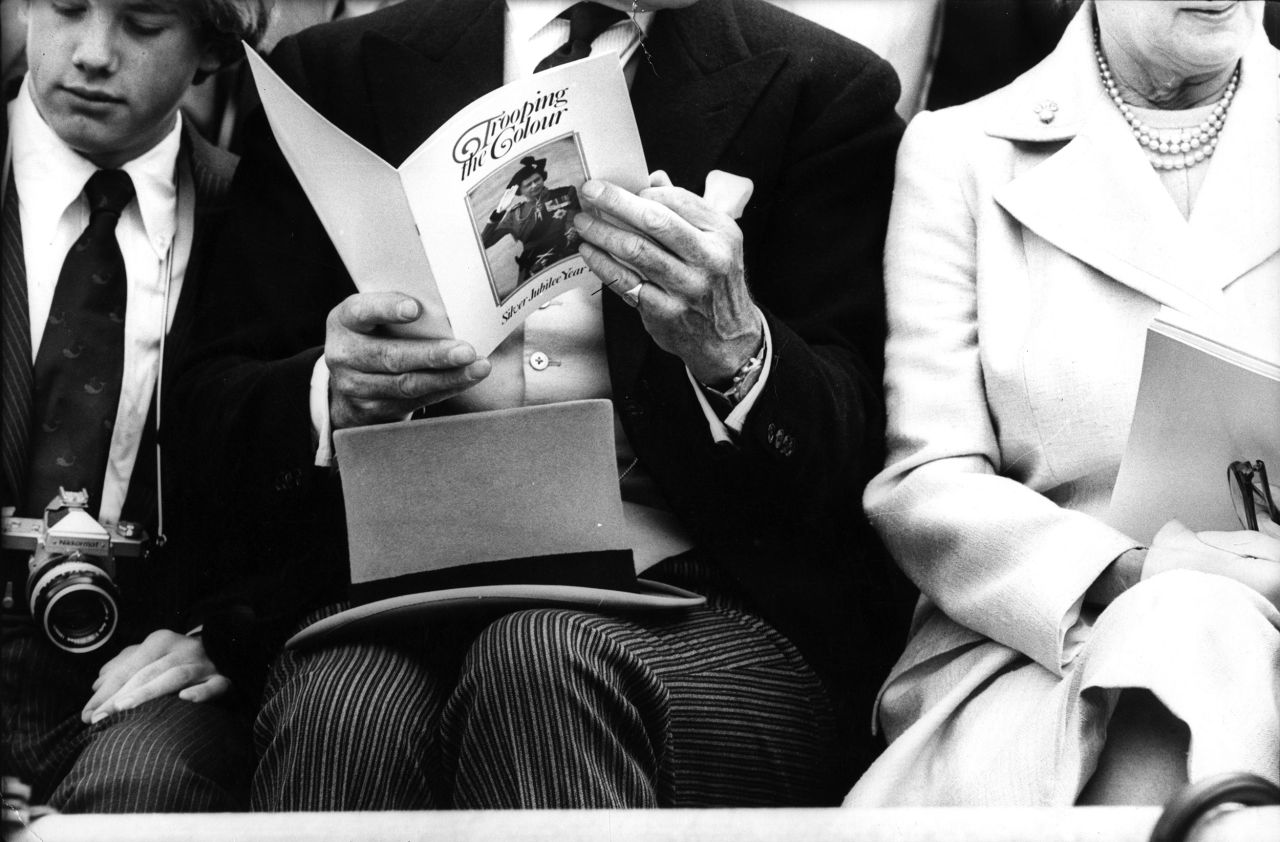 A sharply dressed man in morning suit rests his top hat on his lap as he reads the program for the Silver Jubilee Trooping the Colour ceremony.
