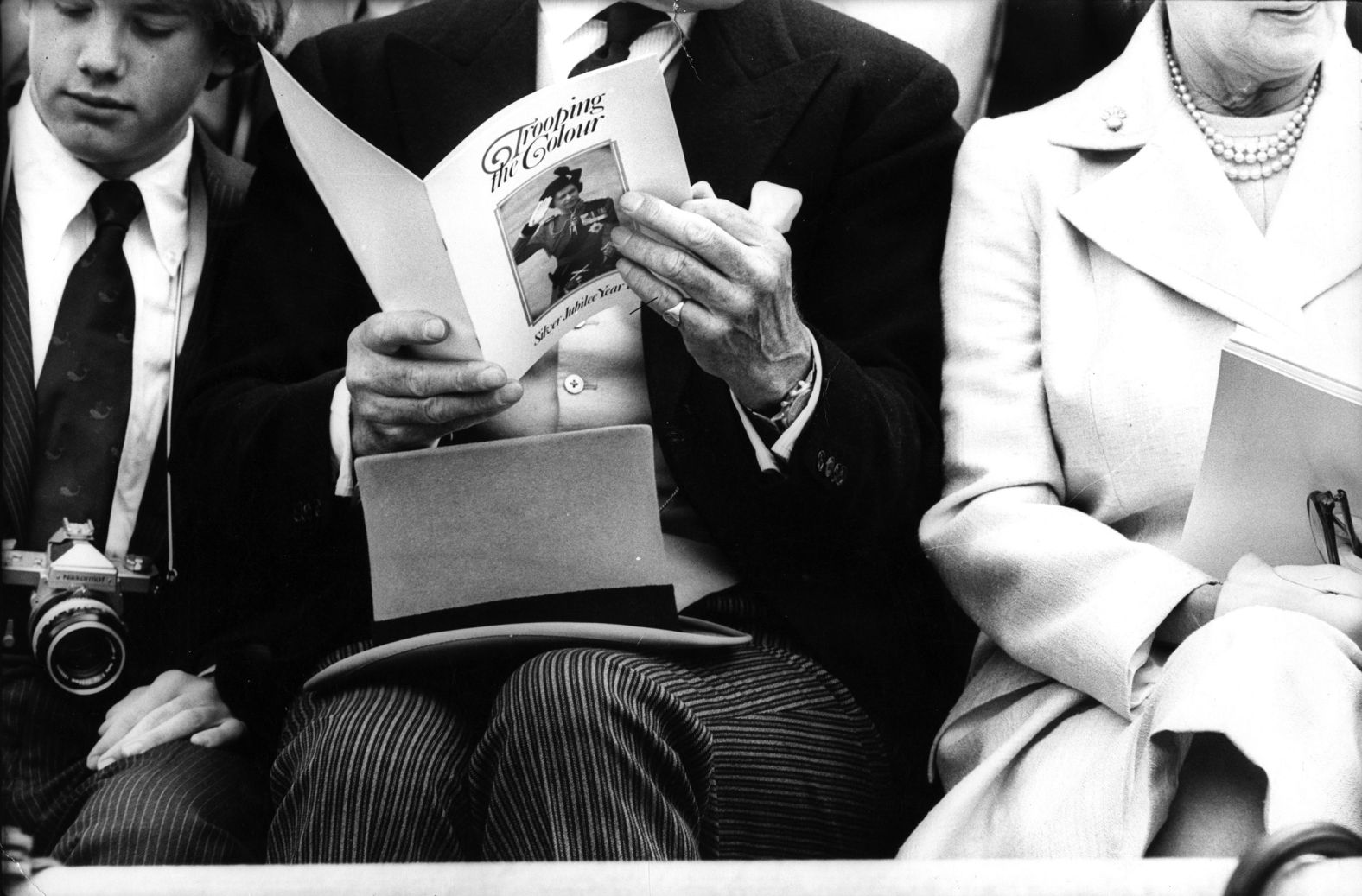 A sharply dressed man in morning suit rests his top hat on his lap as he reads the program for the Silver Jubilee Trooping the Colour ceremony. 