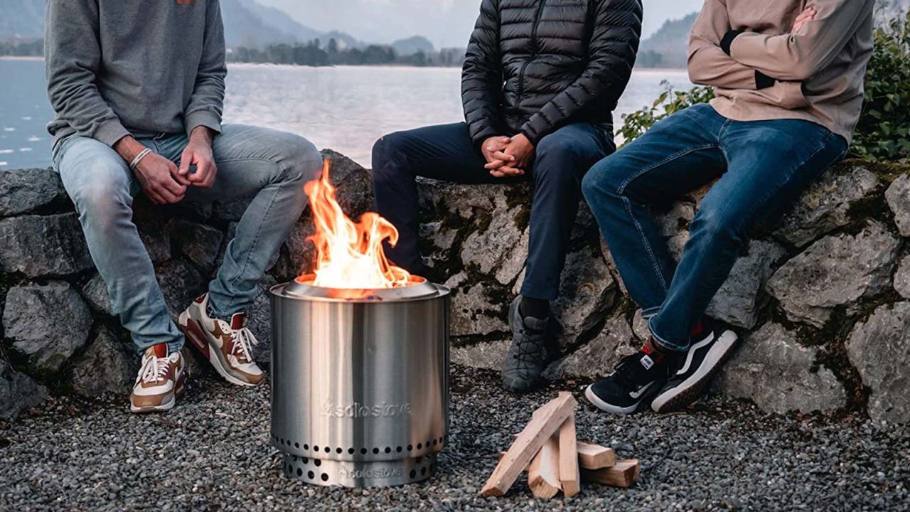 Three people seated by a portable stainless steel fire pit near a rocky shore with firewood on the ground.
