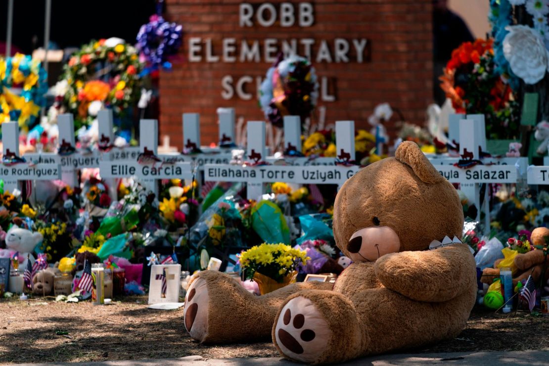 A teddy bear sits in front of crosses with the names of the 21 victims killed at Robb Elementary School.