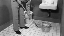 A man wearing a cap and uniform mops the floor of a bathroom at a service station circa 1945.