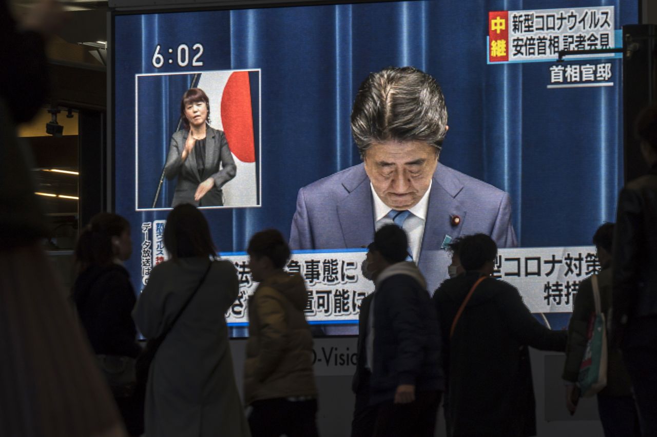 People in Osaka, Japan, walk past a monitor showing a live broadcast of Abe talking about the Covid-19 pandemic in 2020.