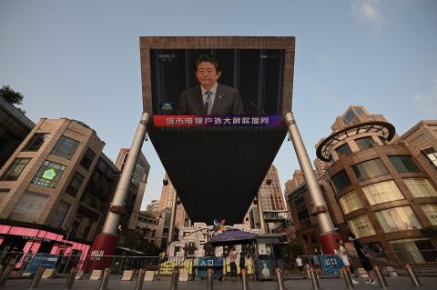 A large video screen shows a news broadcast featuring an image of former Japanese prime minister Shinzo Abe in Beijing.