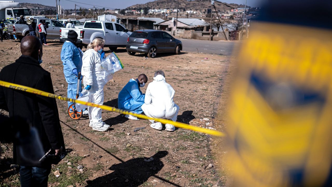 South African Police Service (SAPS) officers enforce a perimeter around a crime scene as pathalogical investigators inspect the crime scene.