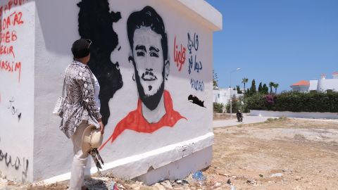 Samia stands by a mural of her son in their hometown of Kelibia. She said his friends, who regularly visit her, painted it. The drawing has his face, the date of his disappearance, a map of Tunisia and says "Fadi always in our hearts."