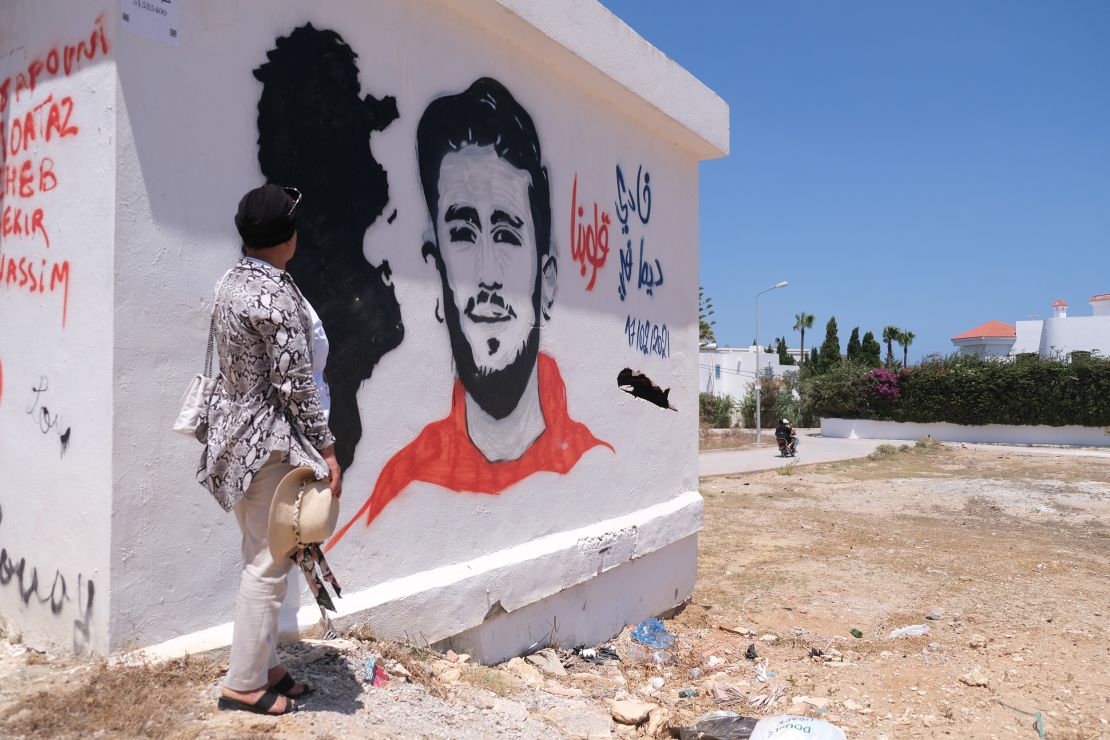 Samia stands by a mural of her son in their hometown of Kelibia. She said his friends, who regularly visit her, painted it. The drawing has his face, the date of his disappearance, a map of Tunisia and says "Fadi always in our hearts."