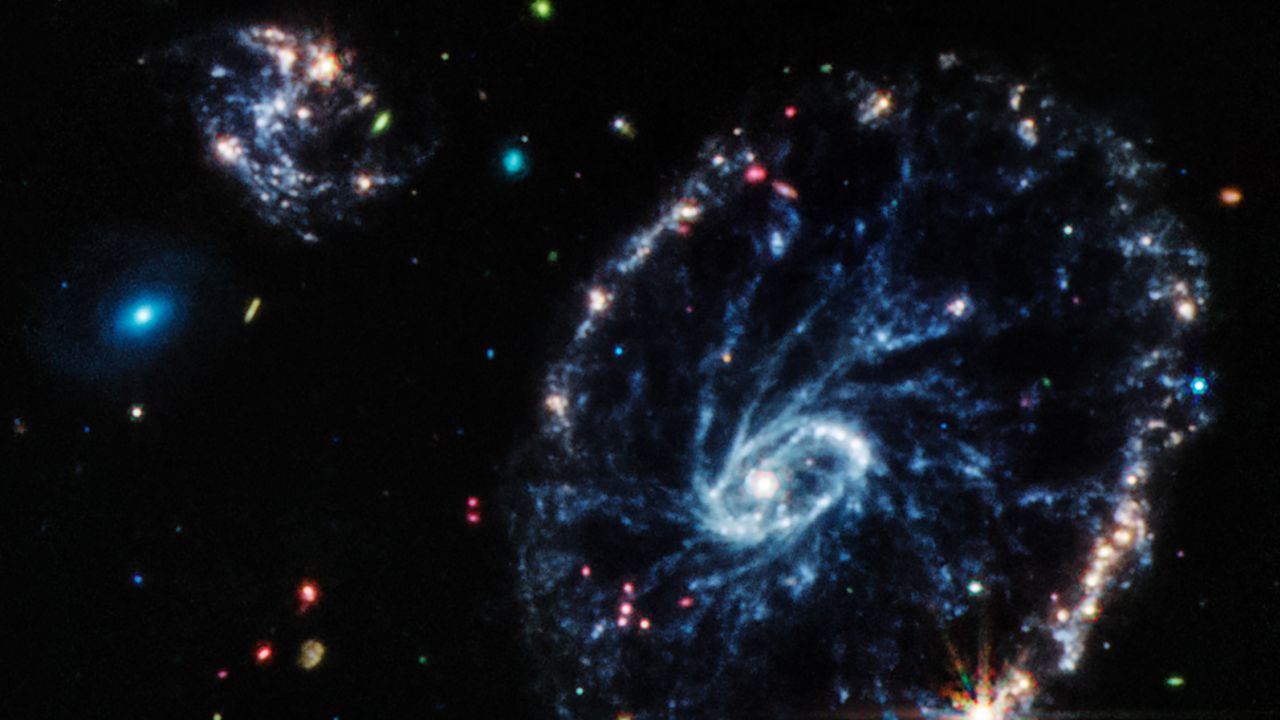 This image from Webb's Mid-Infrared Instrument (MIRI) shows a group of galaxies, including a large distorted ring-shaped galaxy known as the Cartwheel. The Cartwheel Galaxy, located 500 million light-years away in the Sculptor constellation, is composed of a bright inner ring and an active outer ring. While this outer ring has a lot of star formation, the dusty area in between reveals many stars and star clusters. The mid-infrared light captured by MIRI reveals fine details about these dusty regions and young stars within the Cartwheel Galaxy, which are rich in hydrocarbons and other chemical compounds, as well as silicate dust, like much of the dust on Earth. Young stars, many of which are present in the bottom right of the outer ring, energise surrounding hydrocarbon dust, causing it to glow orange. On the other hand, the clearly defined dust between the core and the outer ring, which forms the "spokes" that inspire the galaxy's name, is mostly silicate dust. The smaller spiral galaxy to the upper left of Cartwheel displays much of the same behaviour, showing a large amount of star formation.? MIRI was contributed by ESA and NASA, with the instrument designed and built by a consortium of nationally funded European Institutes (the MIRI European Consortium) in partnership with JPL and the University of Arizona.