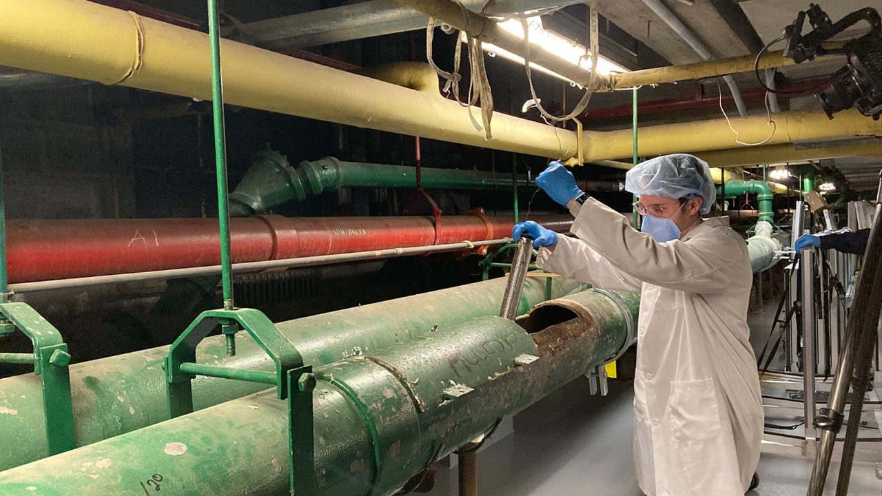 Queens College Research Assistant Justin Silbiger collects a wastewater sample from a sewage pipe in the basement of NYC Health + Hospitals/Elmhurst.