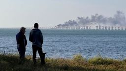 People look at thick black smoke rising from a fire on the Kerch bridge that links Crimea to Russia, after a truck exploded, near Kerch, on October 8, 2022. Moscow announced on October 8, 2022 that a truck exploded igniting a huge fire and damaging the key Kerch bridge -- built as Russia's sole land link with annexed Crimea -- and vowed to find the perpetrators, without immediately blaming Ukraine. 