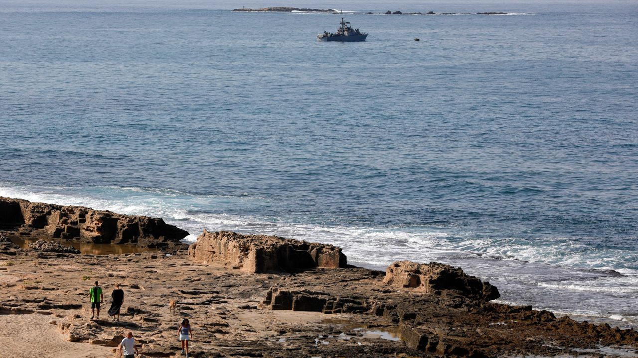 People walk along the beach as a navy vessel patrols the Mediterranean waters off Rosh Hanikra, known in Lebanon as Ras al-Naqura, on the Israeli side of the border between the two countries, on October 7.