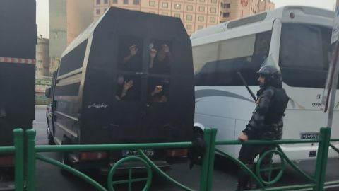 A group of people look out from what appears to be a security van in Tehran, as an officer stands nearby.