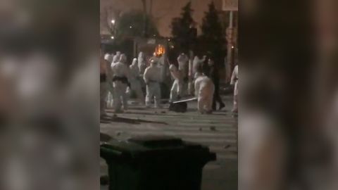 A group of security officers dressed in hazmat suits kick and punch a worker lying on the ground. 