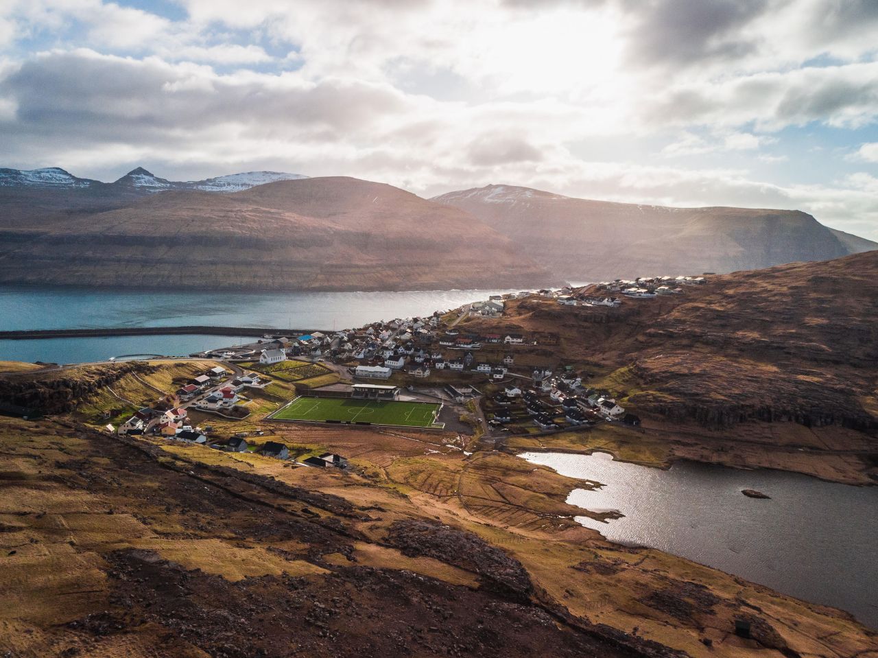 Aerial view of the village Eioi with snow-covered mountain range, football field and dramatic sky during sunset (Faroe Islands, Denmark, Europe)