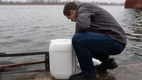 A man pours water into containers from the Dnieper River, and on the other side of the waterway is Russian-controlled territory.