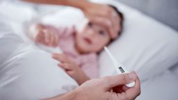 Hands, fever and baby with the hand of a mother taking the temperature with a thermometer of a sick daughter in bed at home. Female parent measuring or testing the symptoms of her girl in the bedroom.