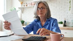 Senior mature business woman holding paper bill using calculator, old lady managing account finance, calculating money budget tax, planning banking loan debt pension payment sit at home kitchen table.