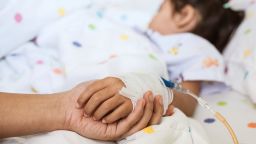 Mother hand holding sick daughter hand who have IV solution bandaged with love and care while she is sleeping on bed in the hospital