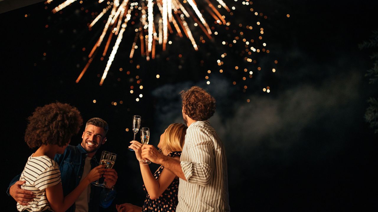 Four friends standing outside, toasting with glasses of wine and watching fireworks