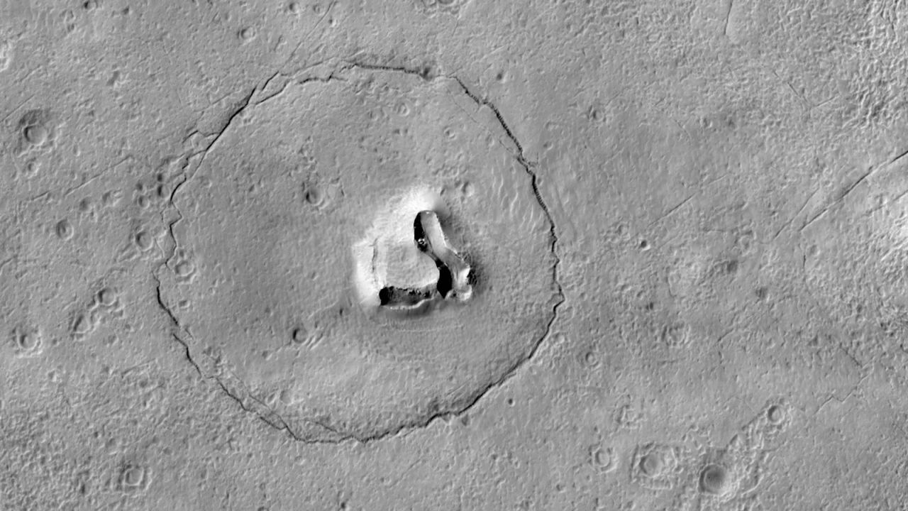 The face of a bear appears to take shape on the Martian surface in this new image taken by the HiRISE camera aboard the Mars Reconnaissance Orbiter. Two craters create the eyes, a circular fracture shapes the face, and a V-shaped collapse structure represents the nose. 