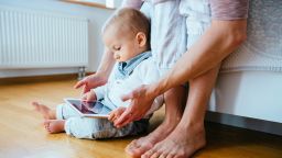 Cute infant baby boy barefoot using a tablet sitting on the flloor, his unrecognizable mother helping him to hold gadget. Real people life concept.