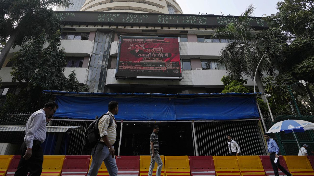 People walk past an electronic display featuring news about Adani Group outside the Bombay Stock Exchange building in Mumbai, India, Friday, Jan. 27, 2023. 