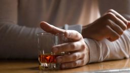 Close up man holding glass with alcohol in hand on wooden table at home, drinking whiskey alone, customer sitting at bar counter, problem with alcoholic beverages concept