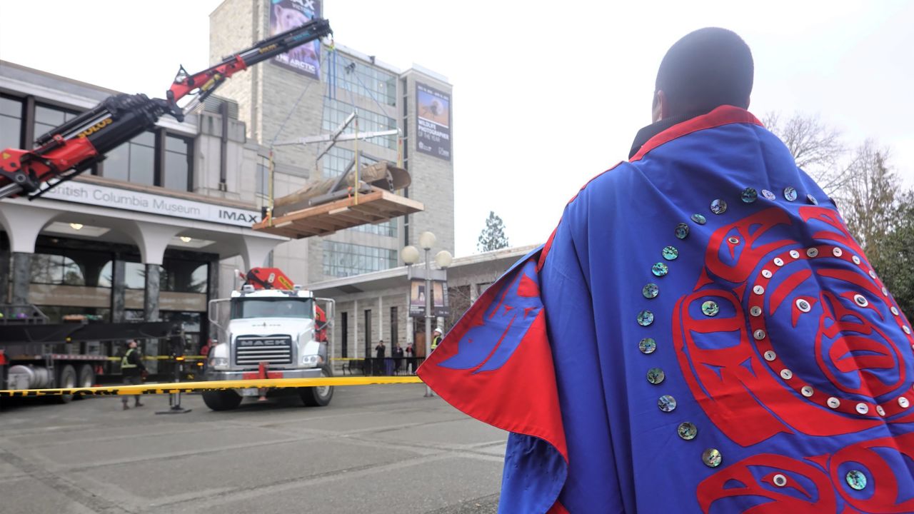 An Indigenous tribal member wearing their clan's regalia, embroidered with their family crest, watches as the totem pole is lifted out of the Royal BC Museum and lowered to the ground.