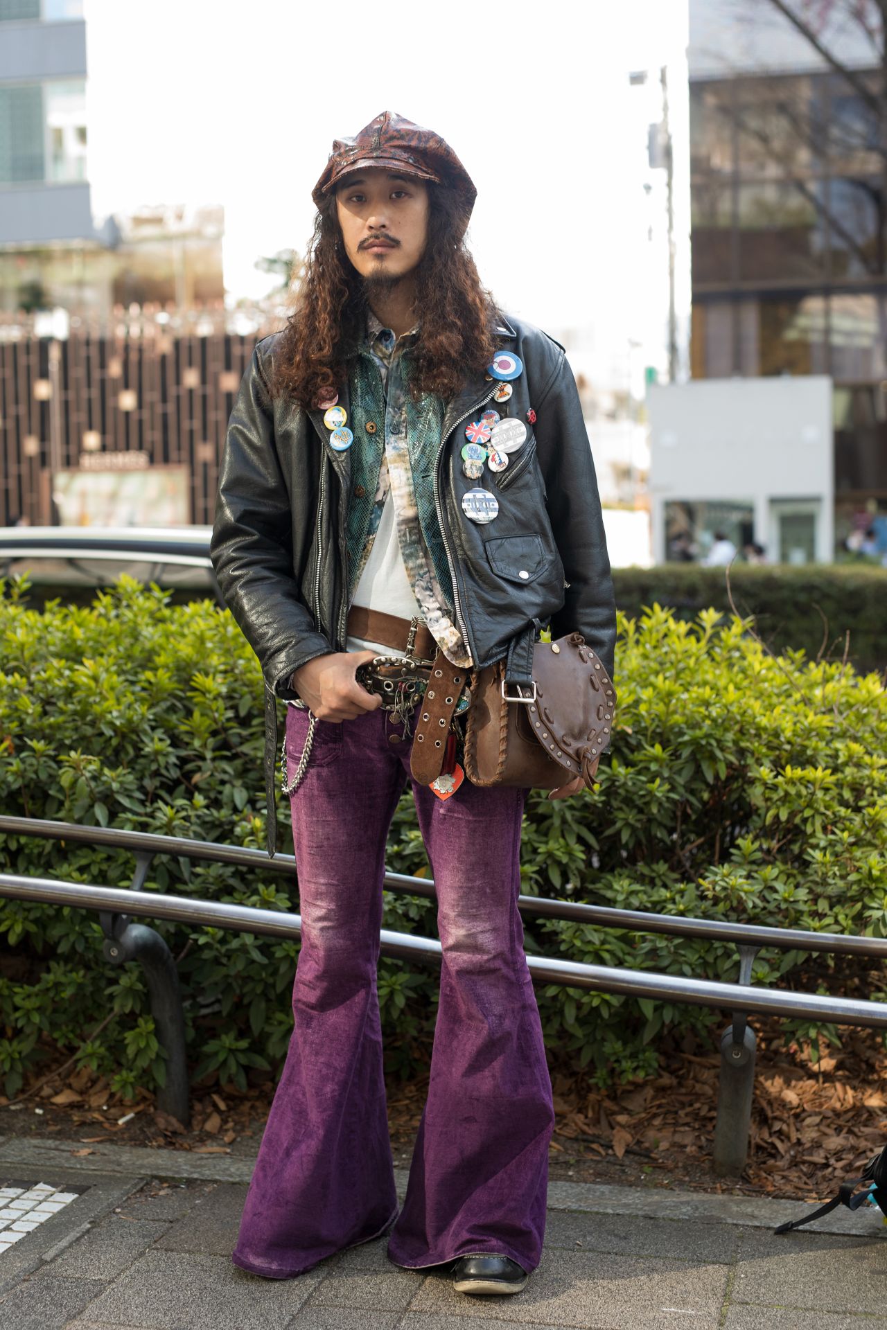 Stood outside the HEoS show, a guest pairs purple flare pants with a black leather jacket while accessorizing with a brown newsboy hat and studded bag.