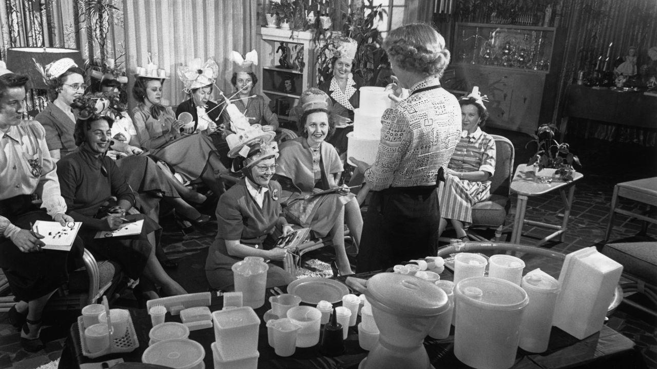 circa 1950:  A woman holds three Tupperware containers while standing in front of a group of women seated in a living room during a Tupperware party. Some of the women wear hats made from the plastic containers. A table in the foreground displays a range of the company's products.