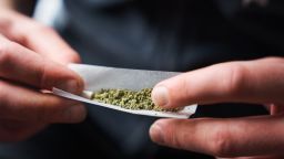 Close-up of a young adult rolling a marijuana joint against a blurred background. Man placing desiccated marijuana leaves inside rolling paper.