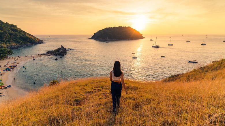 A woman in white t-shirt jeans stands on a mountain with yellow grass overlooking Yanui Beach, the boat and the mountains below. and there is sunlight