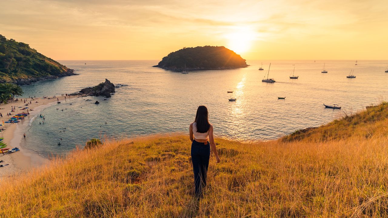 A woman in white t-shirt jeans stands on a mountain with yellow grass overlooking Yanui Beach, the boat and the mountains below. and there is sunlight
