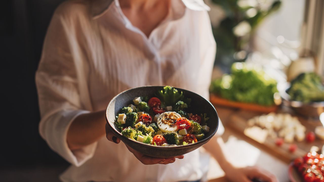 Healthy dinner or lunch. Woman in t-shirt and jeans standing and holding vegan superbowl or Buddha bowl with hummus, vegetable, salad, beans, couscous and avocado and smoothie in hands, square crop