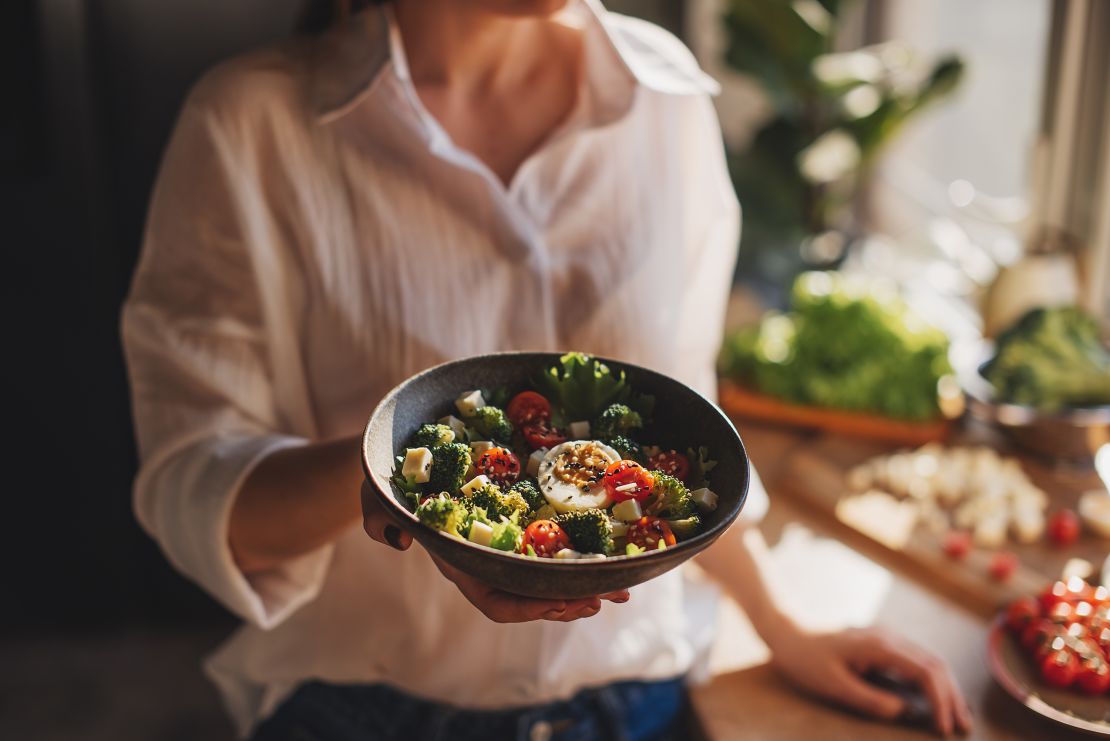 Healthy dinner or lunch. Woman in t-shirt and jeans standing and holding vegan superbowl or Buddha bowl with hummus, vegetable, salad, beans, couscous and avocado and smoothie in hands, square crop
