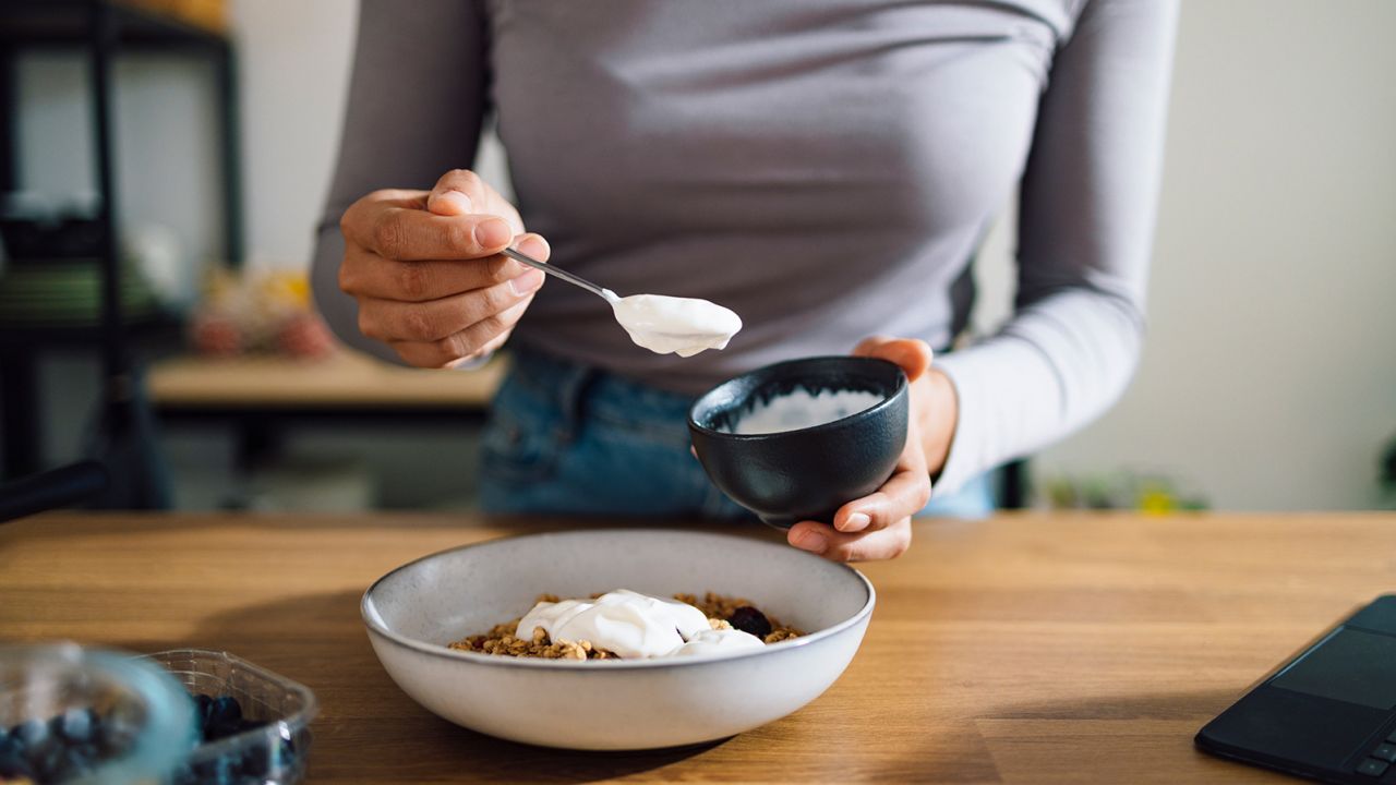 An anonymous Asian woman mixing cereals, greek yogurt and blueberries in a bowl on a kitchen table at home.