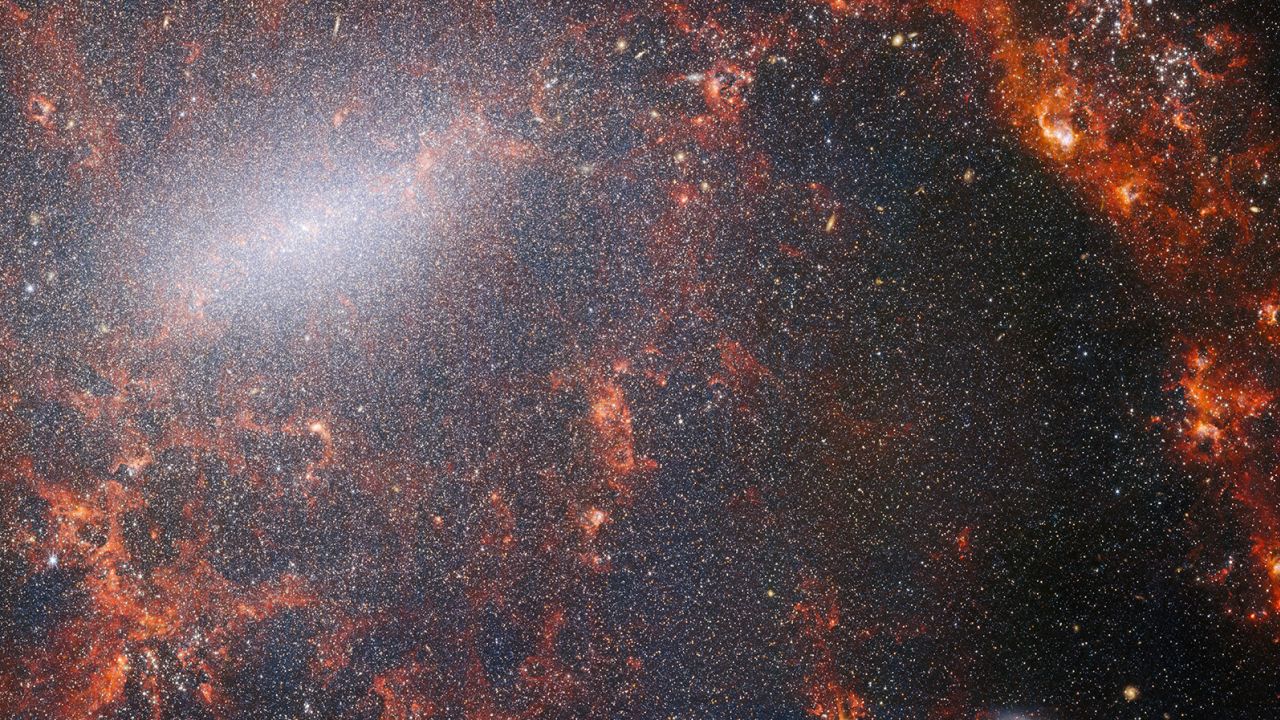 A delicate tracery of dust and bright star clusters threads across this image from the NASA/ESA/CSA James Webb Space Telescope. This view from Webb's NIRCam instrument is studded by the galaxy's massive population of stars, most dense along its bright central bar, along with burning red clouds of gas illuminated by young stars within. These glittering stars belong to the barred spiral galaxy NGC 5068, located around 17 million light-years from Earth in the constellation Virgo. Learn more about this image here. [Image Description: A close-in image of a spiral galaxy, showing its core and part of a spiral arm. At this distance thousands upon thousands of tiny stars that make up the galaxy can be seen. The stars are most dense in a whitish bar that forms the core, and less dense out from that towards the arm. Bright red gas clouds follow the twist of the galaxy and the spiral arm.]