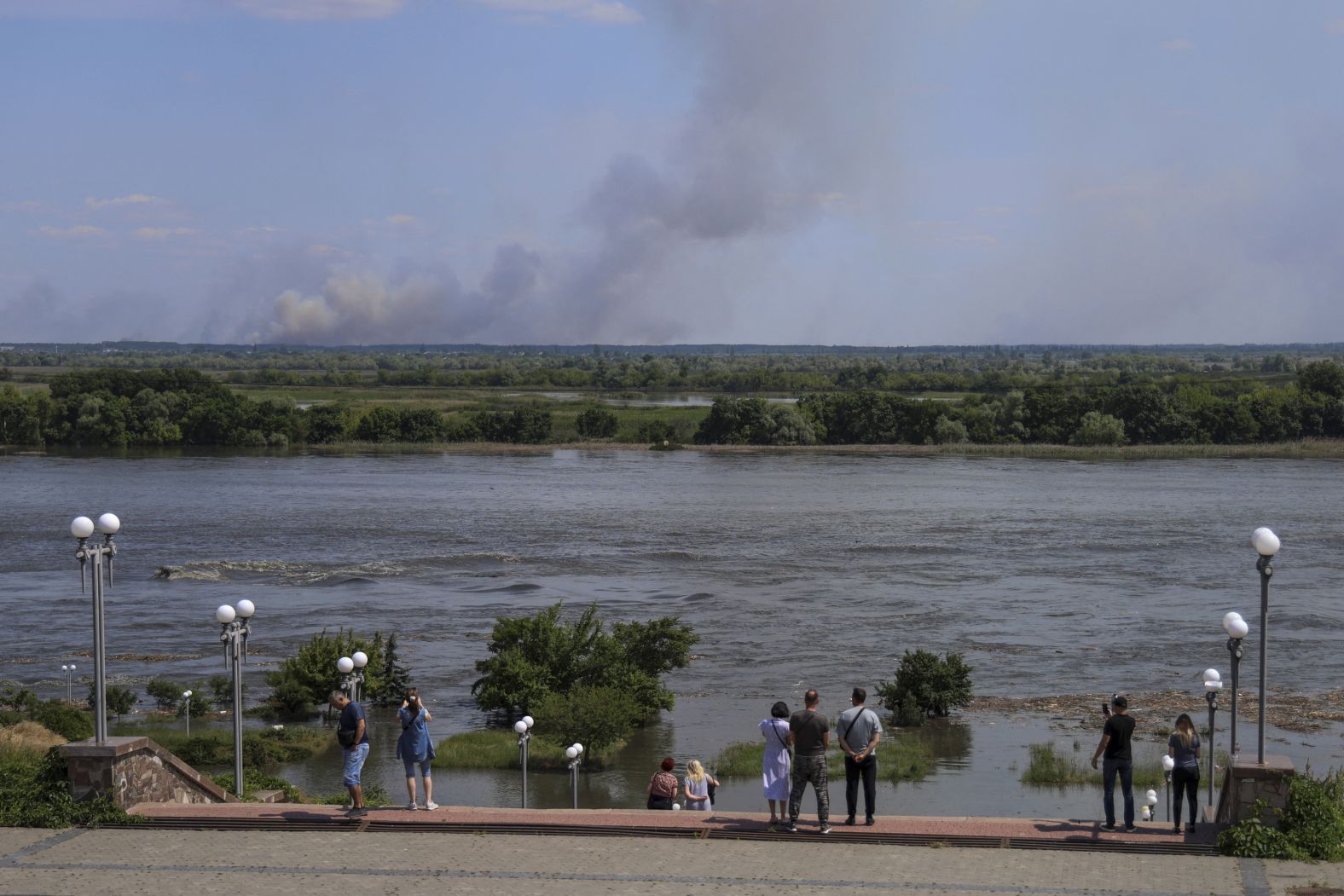 Local residents stand on an embankment of the flooded Dnipro River as smoke rises from shelling on the opposite bank.