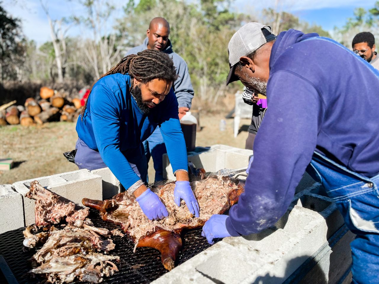 Marvin Ross (left) and Willis Spells pick meat from a roasted hog. Marvin has on blue shirt and his teacher, Spells, has on a cap and a purple sweatshirt with his back to the camera.